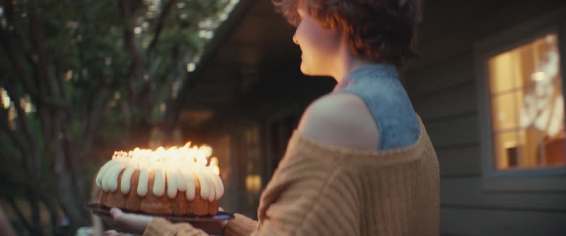 A smiling young woman in a beige cardigan carries a bundt cake with many lit candles toward a table where two other women are seated outside during an evening party