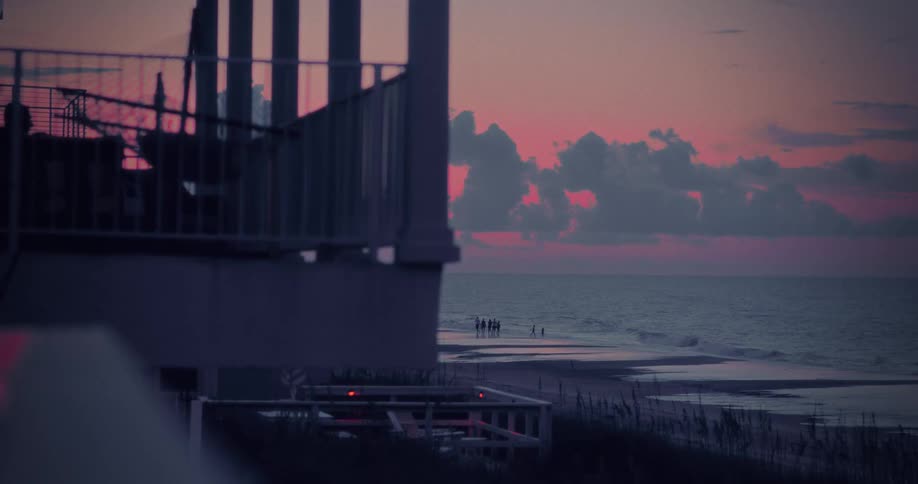 A seaside balcony with a hammock overlooks a quiet beach with a small crowd walking at pink dawn