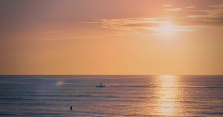 A small boat moves across the dark ocean water at sunset leaving a wake that cuts through the golden sun path while a person rides a jet ski in the water below