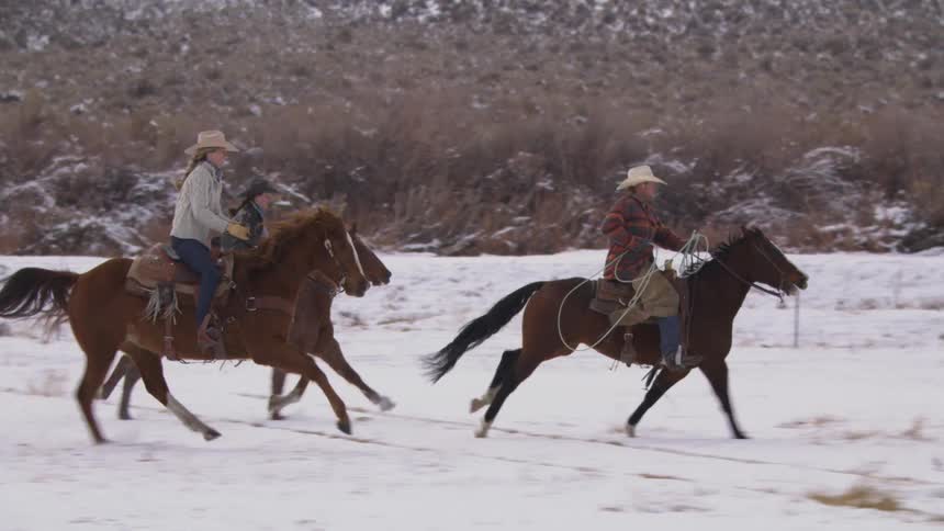 Three people wearing cowboy hats ride horses across a snowcovered landscape at midday