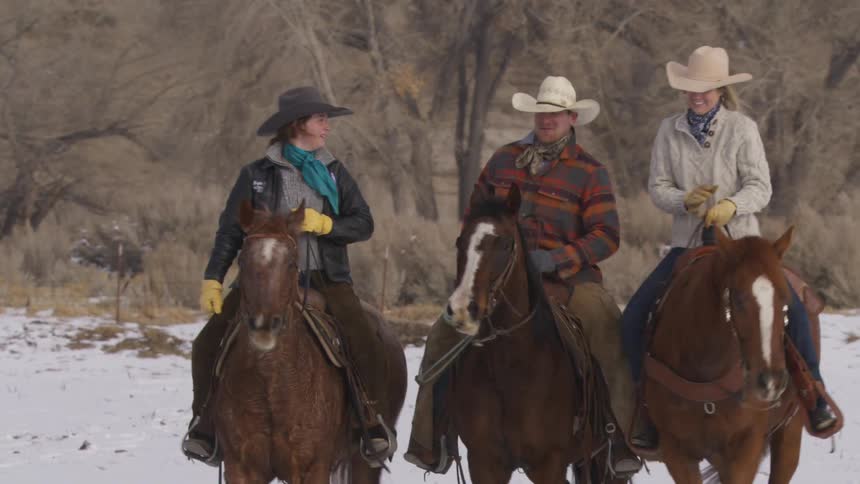 Three people on horseback stand facing the camera in a snowy field the center rider wears a plaid jacket and two riders on the sides wear solid sweaters and they look at each other and smile while riding