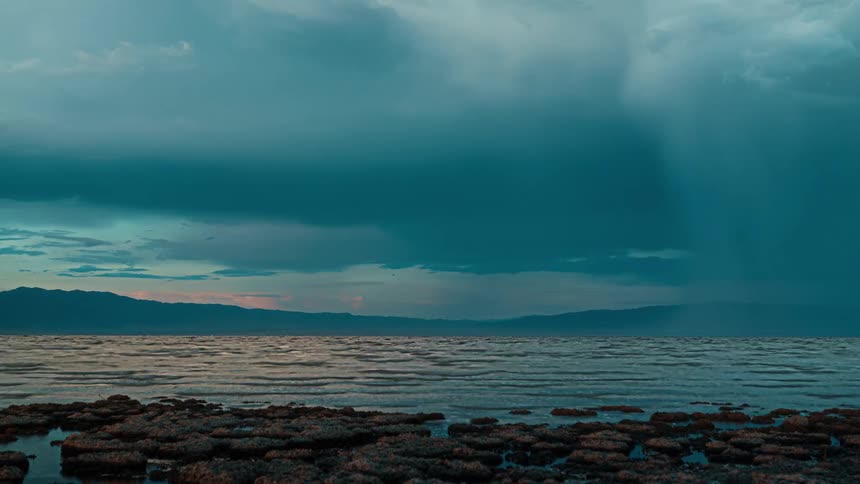 A vast dramatic wide view of a stormy ocean or lake shoreline shows a heavy rain shaft falling from dark blue clouds over distant mountains and a rocky beach in the foreground