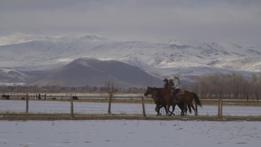 Two people riding horses in a rural setting with snow on the ground and mountains in the background