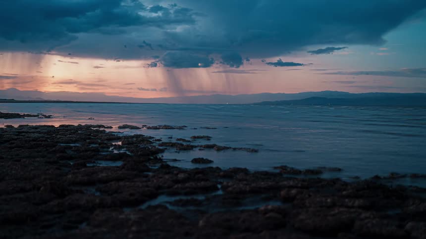 Rainstorm moves across the water near the rocky shore under a red sky