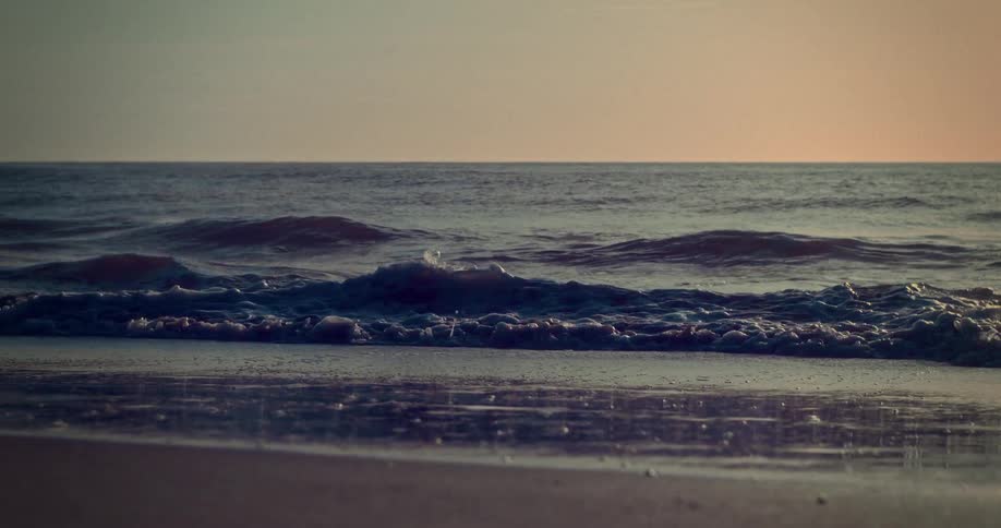 The video is a lowangle shot from the beach capturing a series of waves rolling onto the wet sandthe scene is lit by a soft warm glow from a setting or rising sun on the horizon creating a gentle and tranquil atmosphere with deep blue water tones