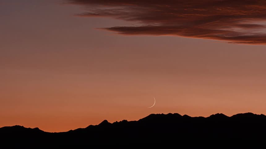 A thin crescent moon hangs above a silhouetted mountain range against a vibrant orange and pink sunset sky with dark clouds