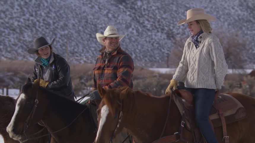 Three riders a man and two women dressed as cowboys and cowgirls in hats and warm clothes sit on their brown horses in a snowy field with mountains in the background