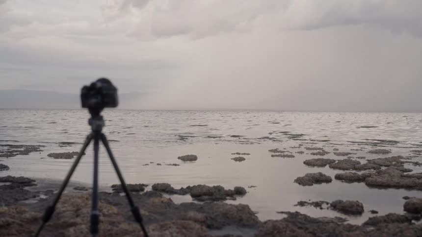 Very small waves are rolling in to a rocky beach where a camera is set up on a tripod