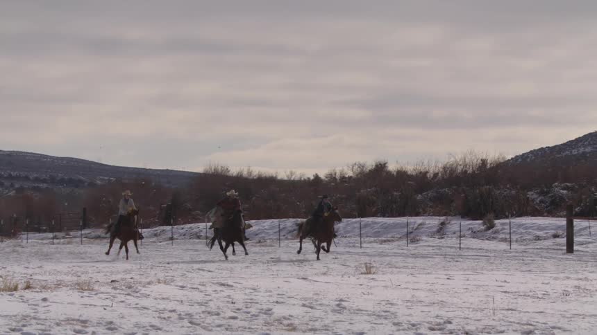 Three people ride horses across a snowcovered ranch at midday