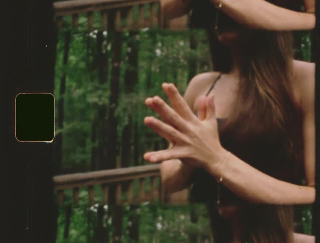 A woman with long brown hair performs a choreographed movement with her hands and arms her face is visible as she looks downward and the background shows a dense forest of tall trees