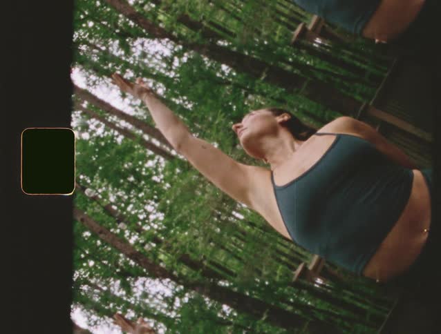 A woman in dark workout clothes performs a stretching pose with her arm extended upward she stands on a wooden structure surrounded by tall trees