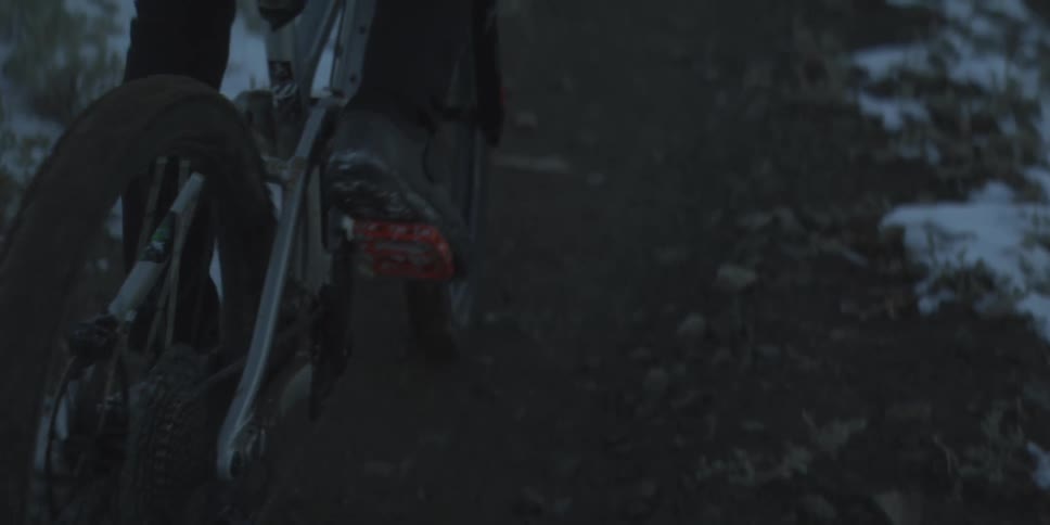 A cyclists foot moves as he pedal a bicycle in a dark lowlight setting the rear wheel and drivetrain are visible and the ground has patches of snow