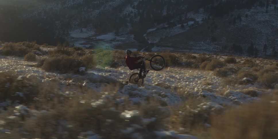 A cyclist on a mountain bike rides across a brushcovered hill then perform a wheelie and a large dark mountain forms the background