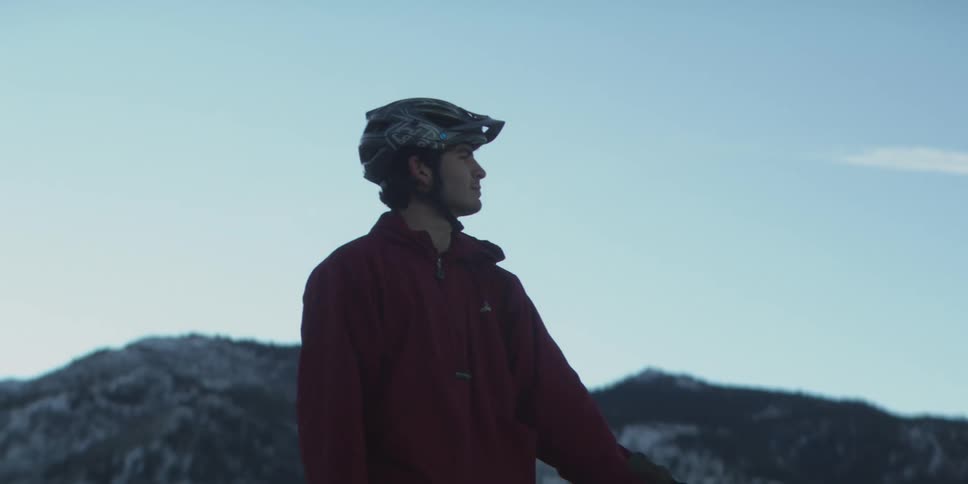 A cyclist wearing a helmet and a red jacket pauses on a snowy mountainous terrain looking off into the distance the shot is a profile silhouette against a bright pale blue sky with a wispy cloud suggesting a highaltitude winter environment