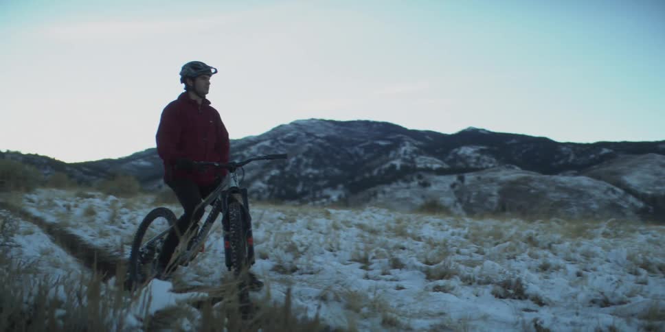 Cyclist in red jacket riding through snowy and grassy terrain with mountains under clear sky