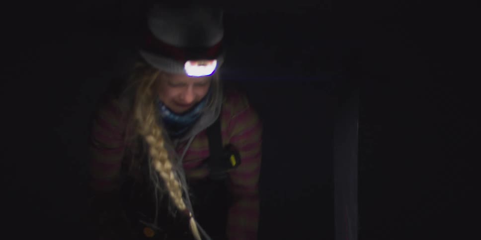 A smiling woman wearing a headlamp and braided hair stands in the dark snow at night searching on a red bag next to an upright ski