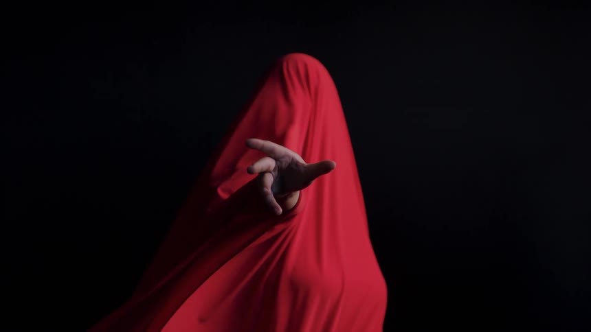 A person draped in a red sheet reaches a hand forward through the fabric against a dark background