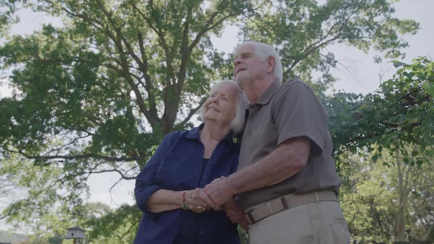 The video is an outdoor lowangle shot of an elderly couple standing close together and holding hands looking up and to the side they are framed against a bright sky and a large lush green tree conveying a sense of peace or wonder