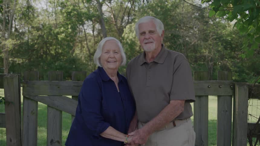 Elderly couple smiles holding hands in outdoor area with wooden fence and surrounding greenery