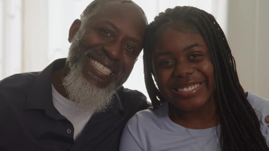 The video is a warm closeup of an older man and a young woman sitting sidebyside sharing a genuine laugh before both turning to smile widely at the camera the man has a distinctive saltandpepper beard and they are sitting in a brightly lit indoor s