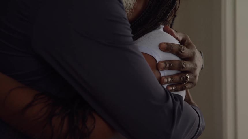The video is a tight closeup focusing on a close embrace between two people showing only their chests and shoulders the person on the right is wearing a lightcolored shirt and is being held by the person on the left who has a darker sleeve and a ring
