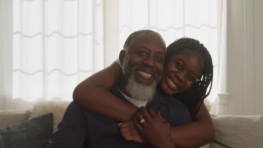 The video is a medium closeup of a smiling african american man with a gray beard and a young woman with braids sitting together on a couch the woman is happily hugging the man from behind with her arms wrapped around his neck and shoulder as they both