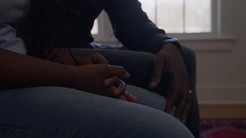 A slow zoom pushes in on a womans clasped hands resting on her lap as she sits closely beside a man on a couch in a sunlit room