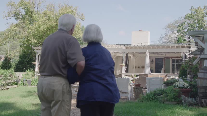 The video is an exterior shot of a senior caucasian couple walking away from the camera on a grassy path in a lush green yard the man has his arm around the womans shoulder as they walk towards a porch or patio area of a house in the background