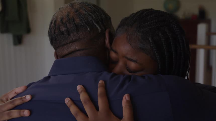 The video shows an african american man with a gray beard and a young woman with braids in a domestic setting they are facing each other and the man pulls the upsetlooking woman into a comforting embrace