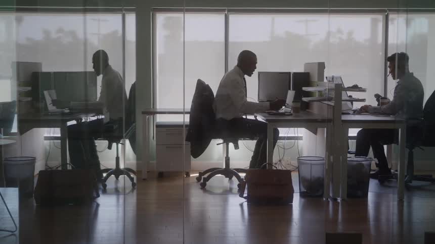 Two men in an office each sitting at their own desk and chair one of the men is talking to the other