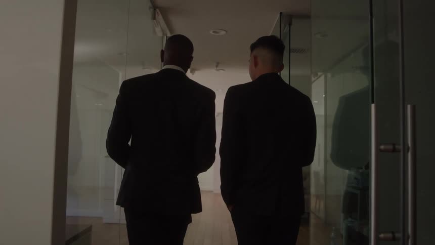 A pair of men in suits walk side by side carrying coffee cups down a modern office hallway seen from behind