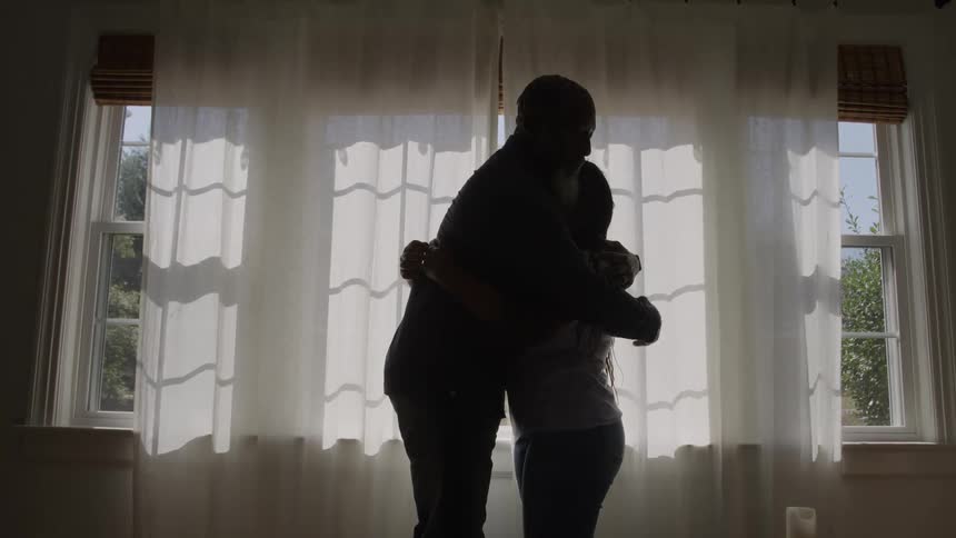 The video shows an african american man and a young woman standing and facing each other becoming silhouetted against two brightly lit windows with sheer curtains the two figures embrace in a warm long hug in the center of the frame