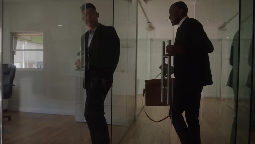 A man with a briefcase holds open a glass door as his colleague with a coffee cup enters the office and he then follows him inside