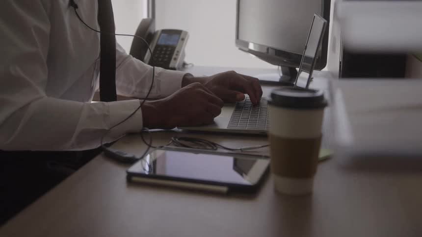 A person works on a laptop at an office desk with a tablet and coffee cup nearby