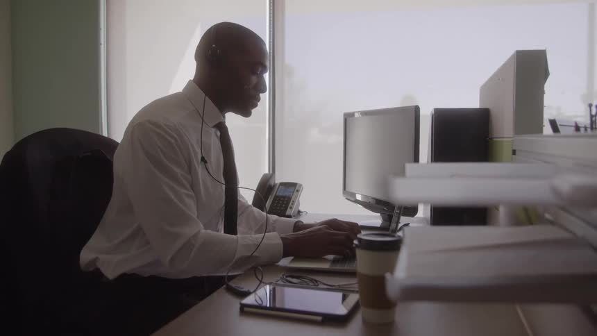 Man wearing a headset types on a laptop while sitting in his office