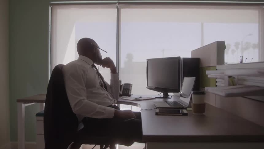 The video shows an african american man in a white dress shirt and dark tie sitting at an office desk with his hand to his chin seemingly deep in thought or on a call he eventually removes his headset and adjusts his tie with the bright light from lar