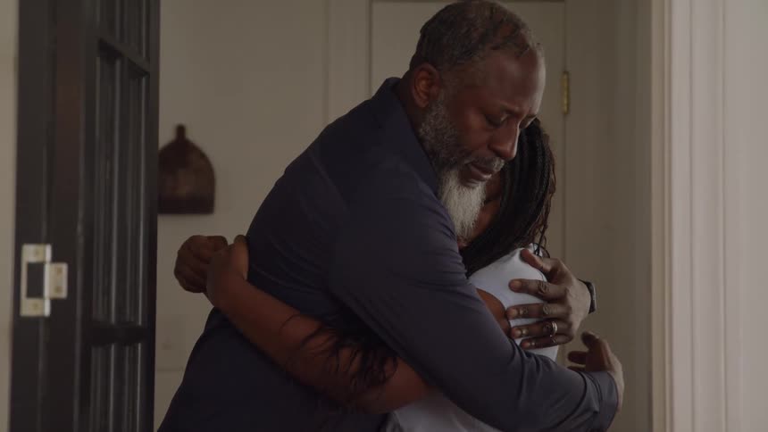 The video shows an african american man with a gray beard comforting a young woman with long braids in a hallway or doorway the woman starts with her arms crossed looking upset and is then pulled into a close tender embrace by the man