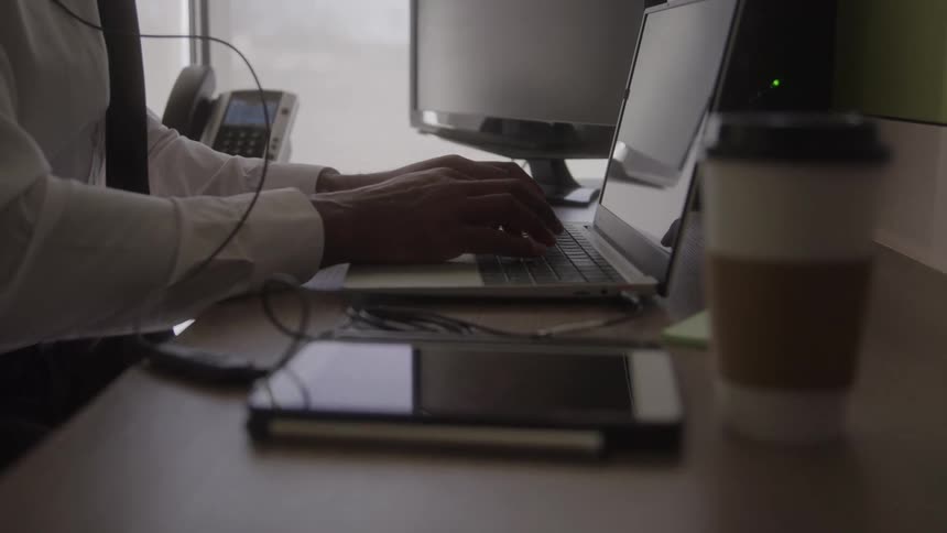 A person in a white shirt types on a laptop at a desk with monitors a phone a tablet and a takeout coffee
