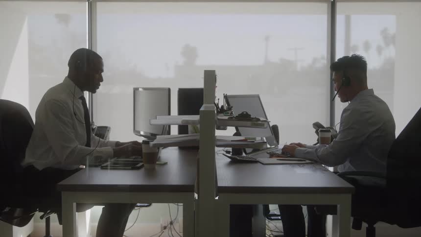 Men wearing headsets work at opposite desks in a bright office
