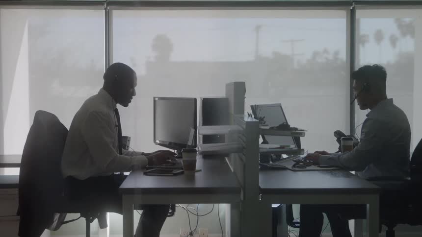 Two individuals work at opposite desks with partition each using computer and headset