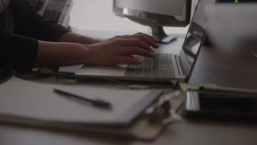 Person types on laptop at desk with clipboard pen monitor and mobile device