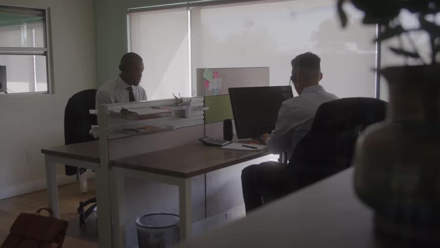 The video is an interior shot of two men in office attire one african american and one caucasian working at separate desks facing away from each other the bright natural light coming through the large background windows creates a soft bright atmospher