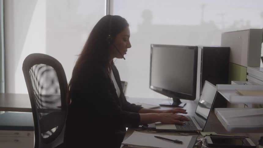 The video is a medium shot of a professional woman with dark hair in a dark blazer and a headset she is seated at a desk typing on a laptop with computer monitors and a bright window in the background