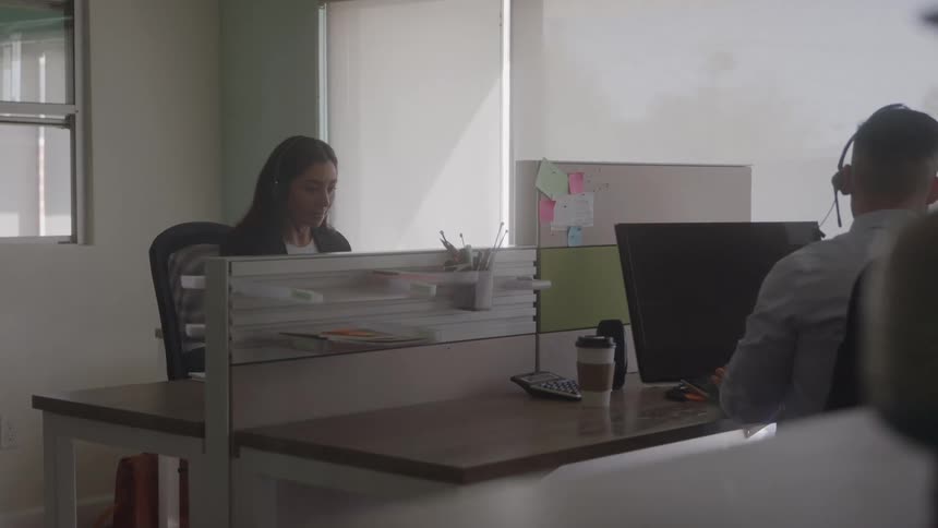 Two individuals work at desks separated by transparent partition with computers calculator coffee cup and window in the background