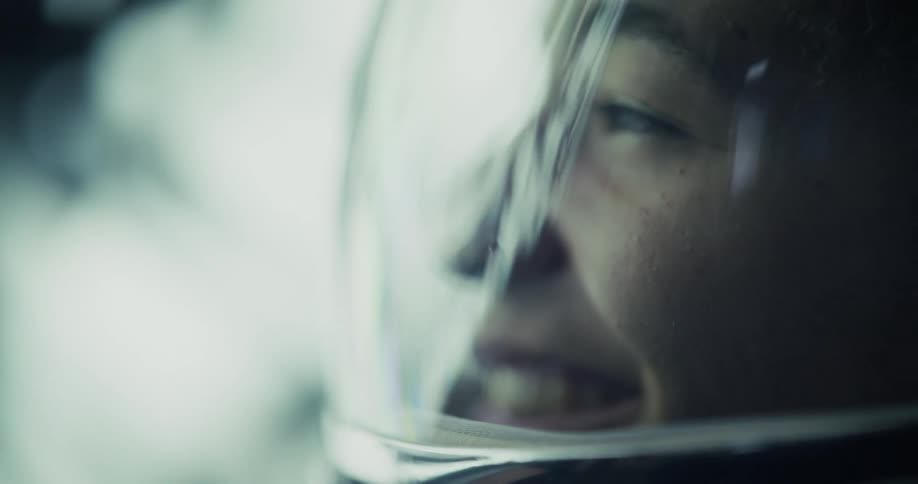 A close profile view shows a womans face reflected in the clear visor of a space helmet in a softly lit dramatic interior setting