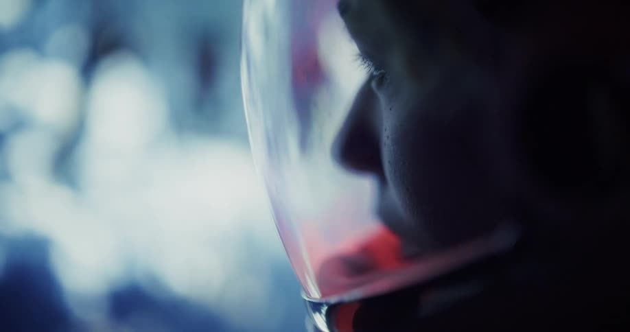 A close profile view shows a womans face reflected in the clear visor of a white helmet under cool blue and red ambient light in a dramatic interior setting