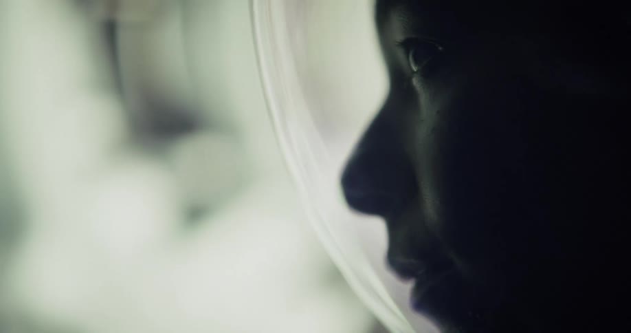 A close up profile shot of a face inside a space helmet looking out onto a brightly lit ambiguous background