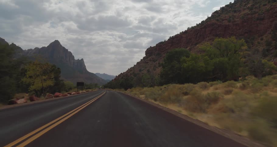 The video is a drivers perspective shot traveling along a winding road through a mountainous area tall reddishbrown rock formations line the road under a cloudy overcast sky giving the scene a dramatic natural feel