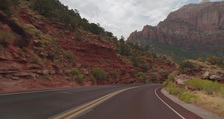 A vehicle view drives along a winding two lane road surrounded by towering red rock cliffs and slopes covered in green brush under a bright overcast sky