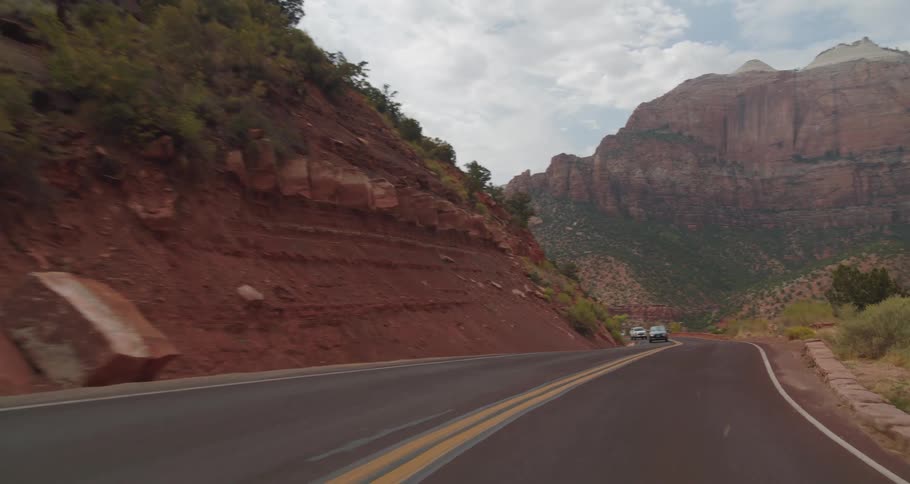 A car winds its way through a scenic mountain pass surrounded by dramatic red rock formations and lush greenery under a cloudy sky